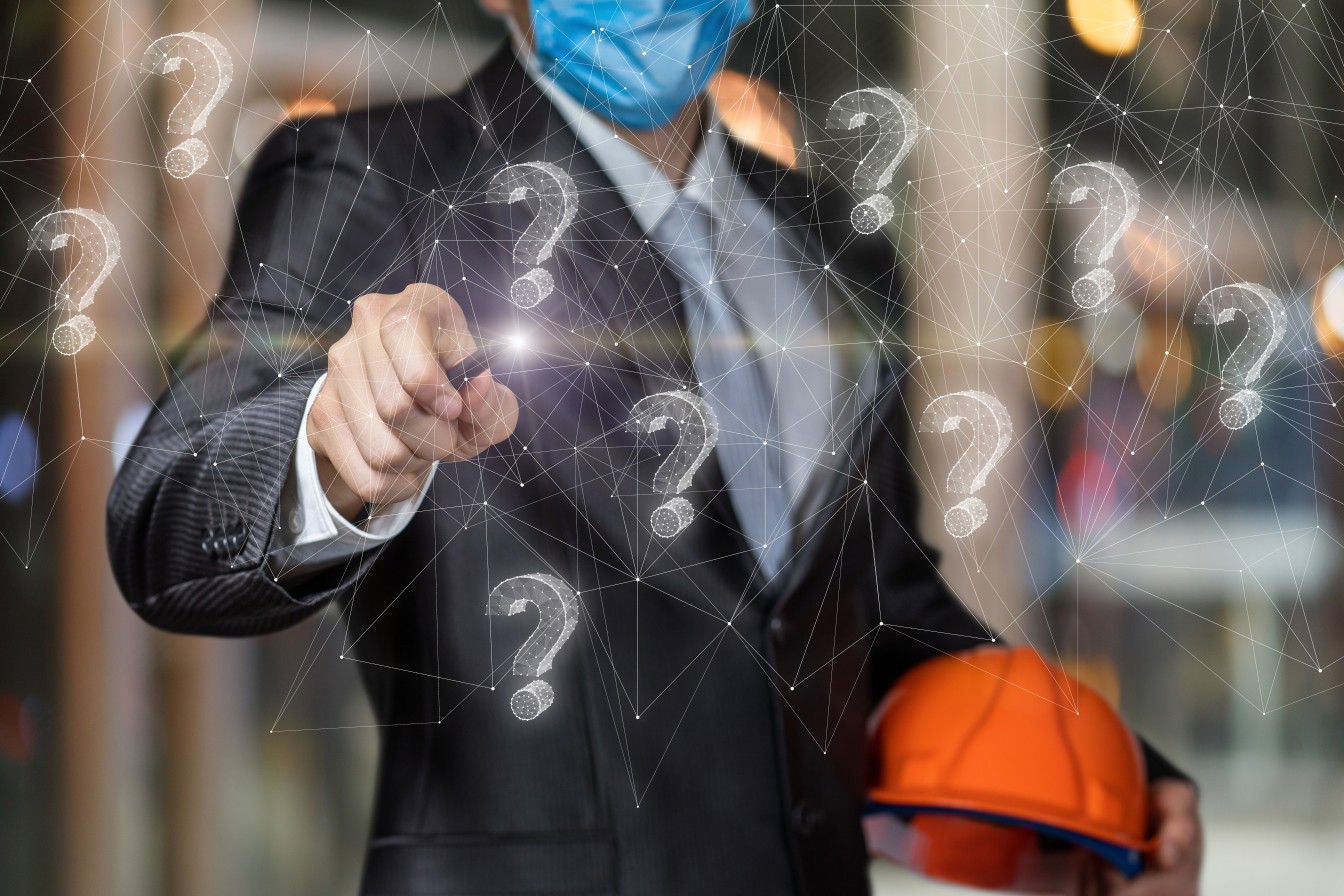 A businessman wearing a protective mask and hardhat, pointing towards question mark icons.